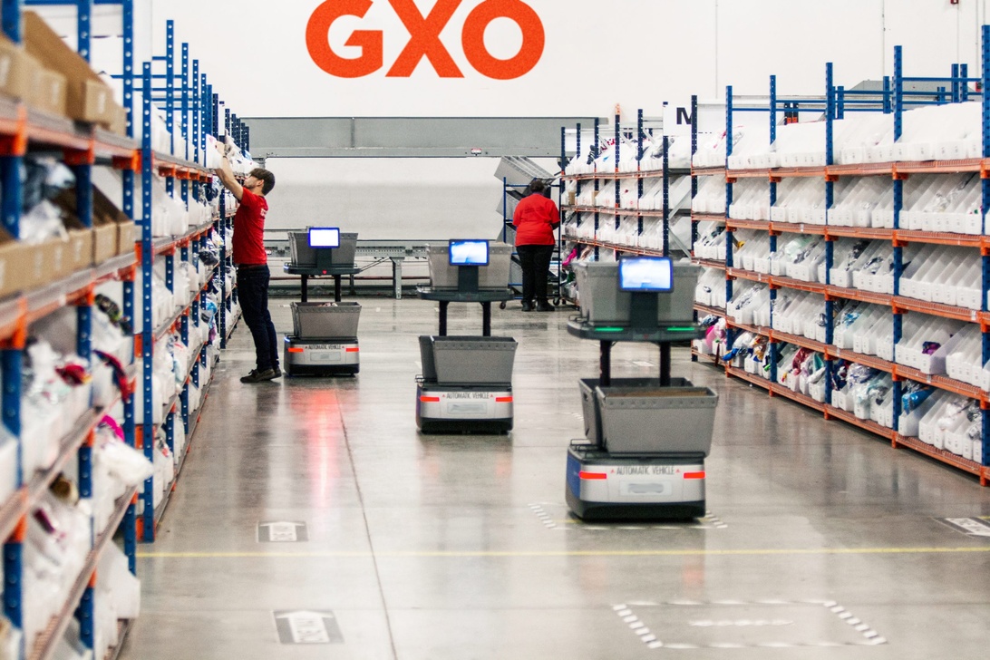 Moderne Lagerhalle mit GXO-Logo, Mitarbeitern in roten Hemden und automatisierten mobilen Robotern auf dem Boden.
